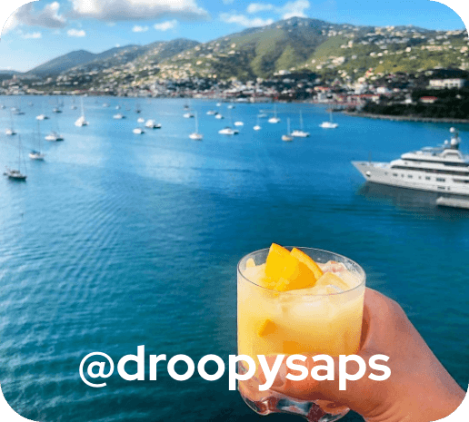 Person holding yellow  tropical drink with water and boats in the background. @droopysaps .